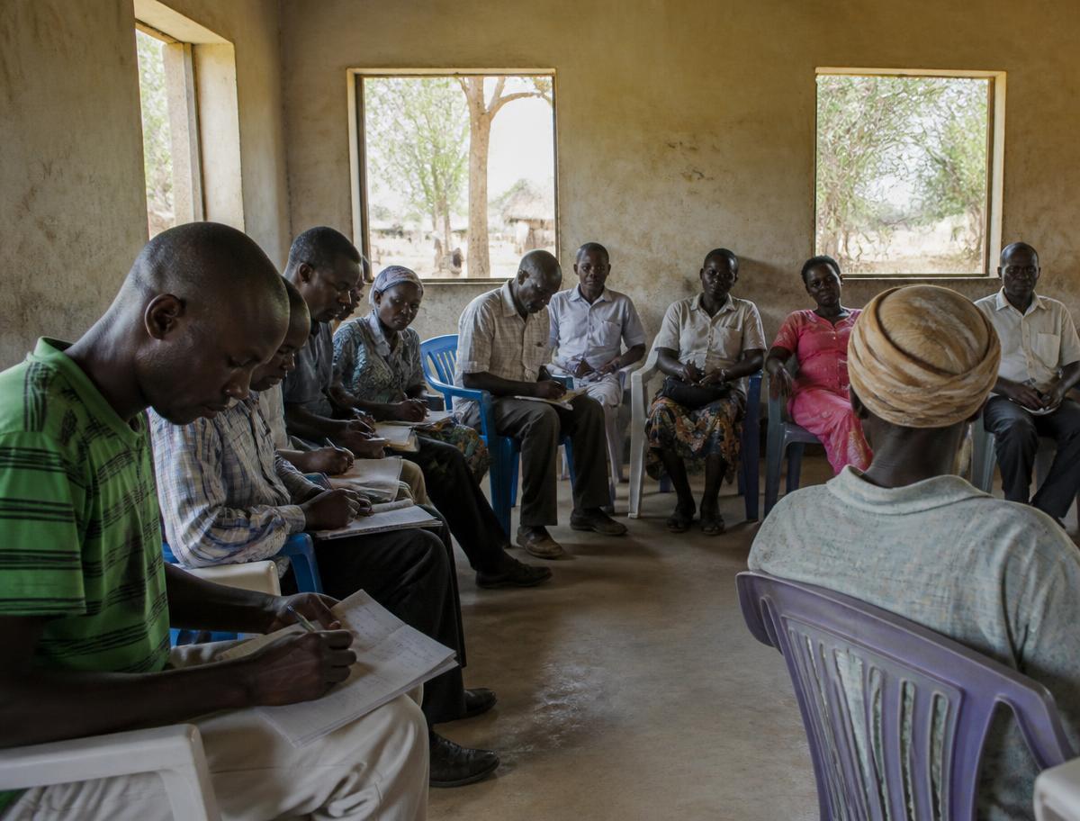 A community meeting in a village hall, participants seated in a semicircle taking notes